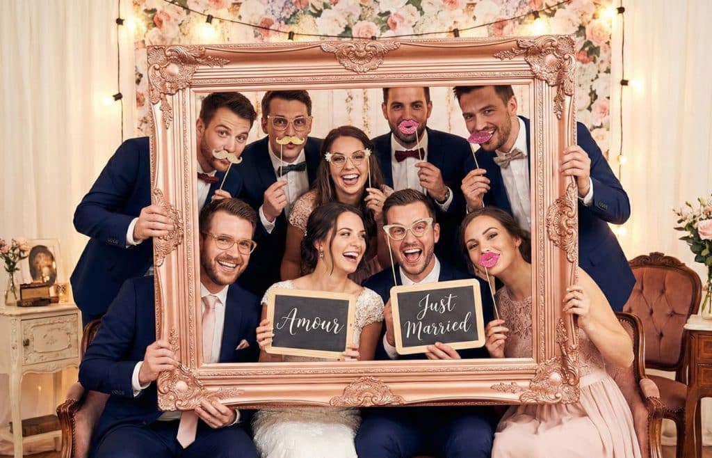 Groupe d'invités de mariage s'amusant avec des accessoires photobooth sur tiges et des pancartes devant un mur de fleurs.