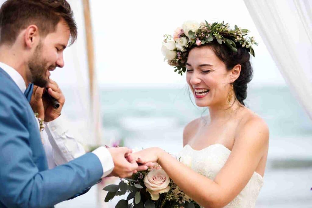 cérémonie de mariage le marié en costume bleu passe la bague à sa femme face à la mer bouquet de fleur couronne sur la tête composition florale
