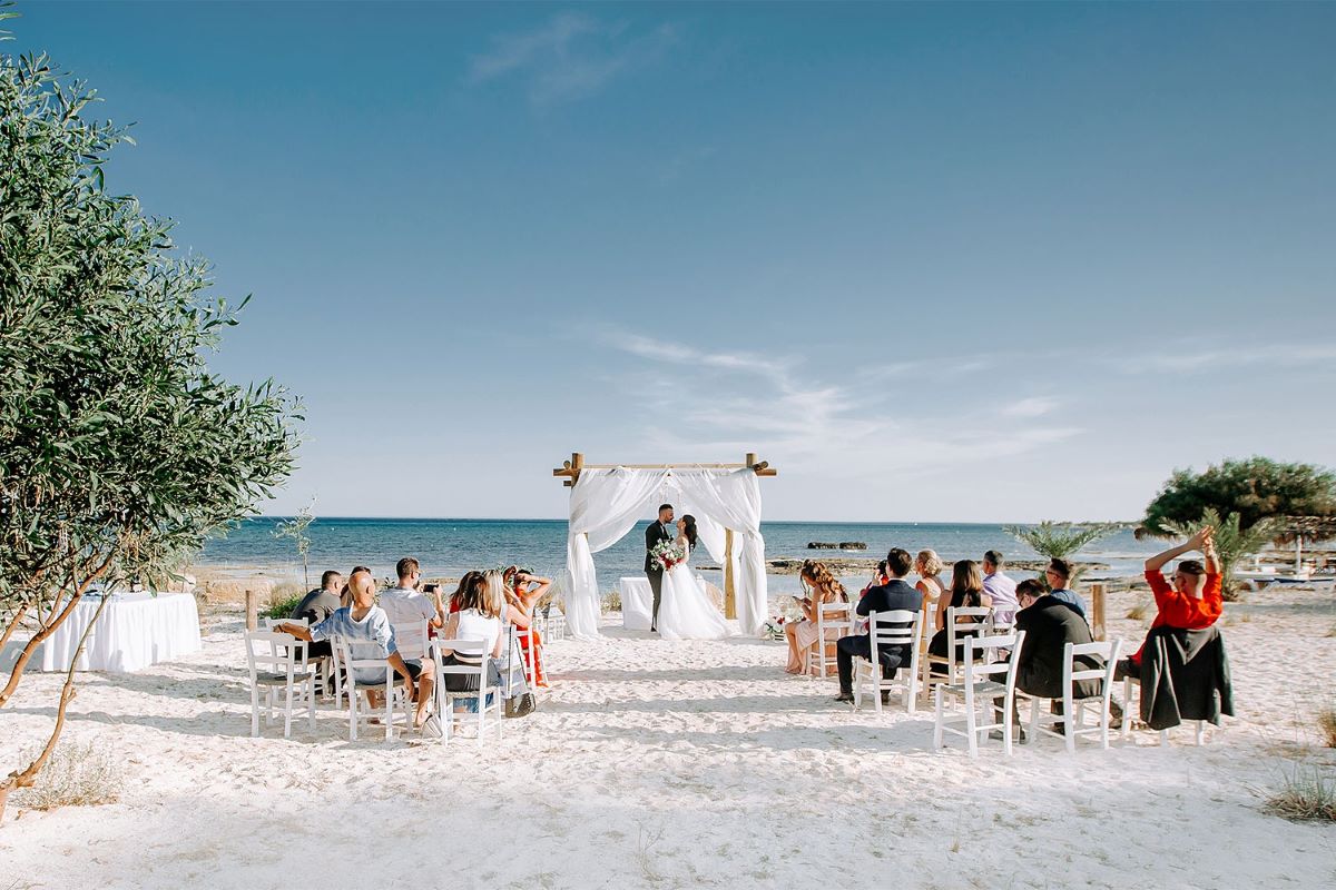 mariage à la plage invités échange des voeux arche en bois drapée blanc chaises sur le sable face à la mer