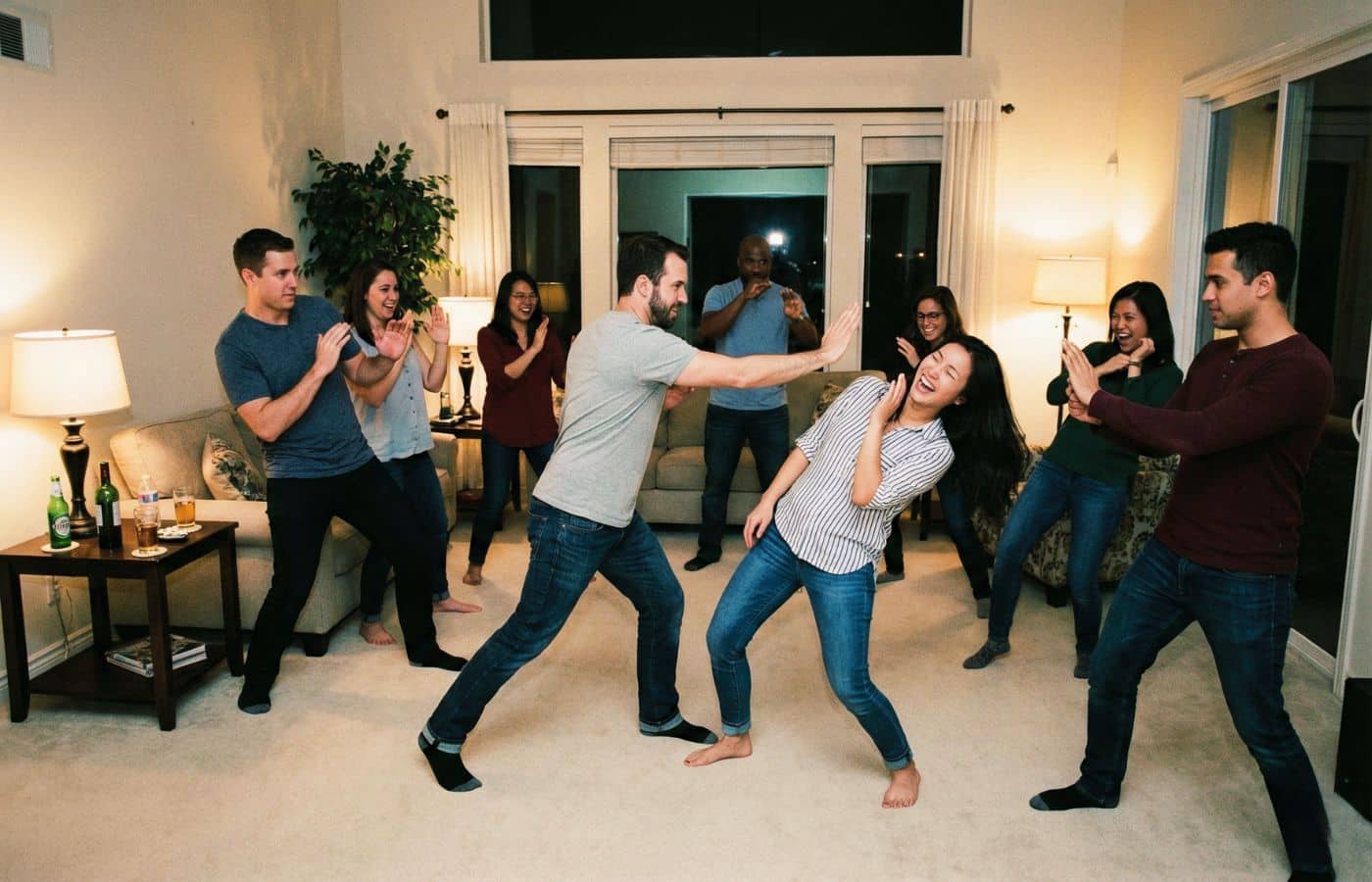 Groupe d'amis adultes jouant au jeu du Ninja dans un salon, prenant des poses d'arts martiaux en riant.