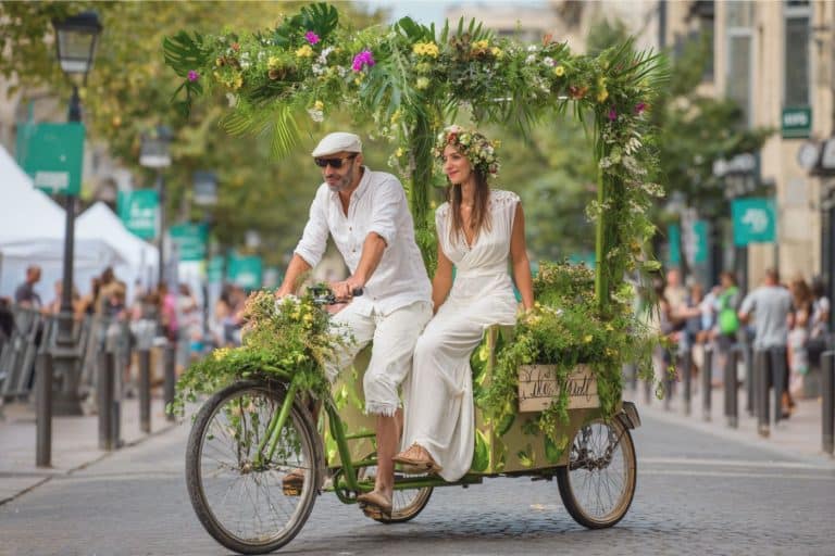 Jeune couple de mariés partant à vélo cargo décoré de fleurs locales, symbole de mobilité douce et d’un mariage éco-responsable et joyeux arrivée mariés à la mairie