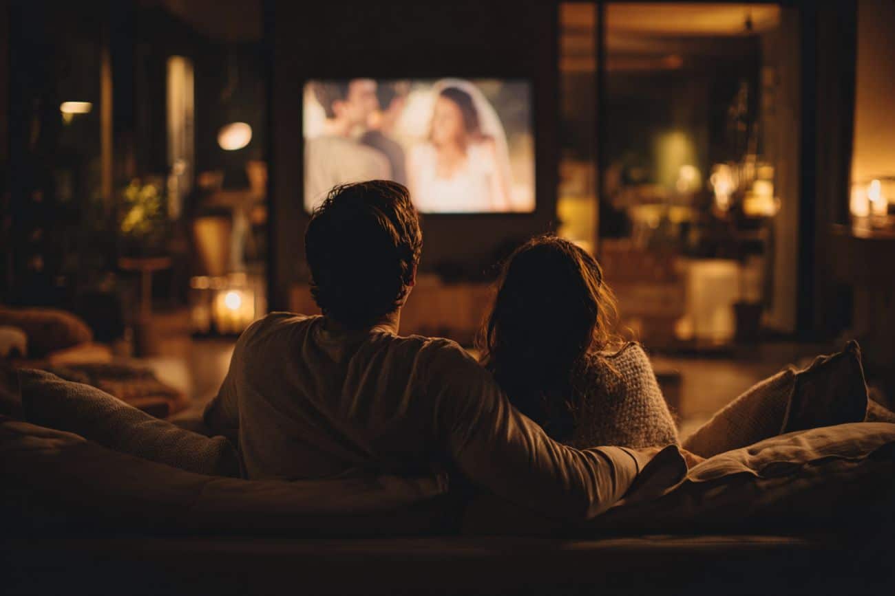 Couple de mariés regardant leur film de mariage sur grand écran dans leur salon, moment émotion et souvenir