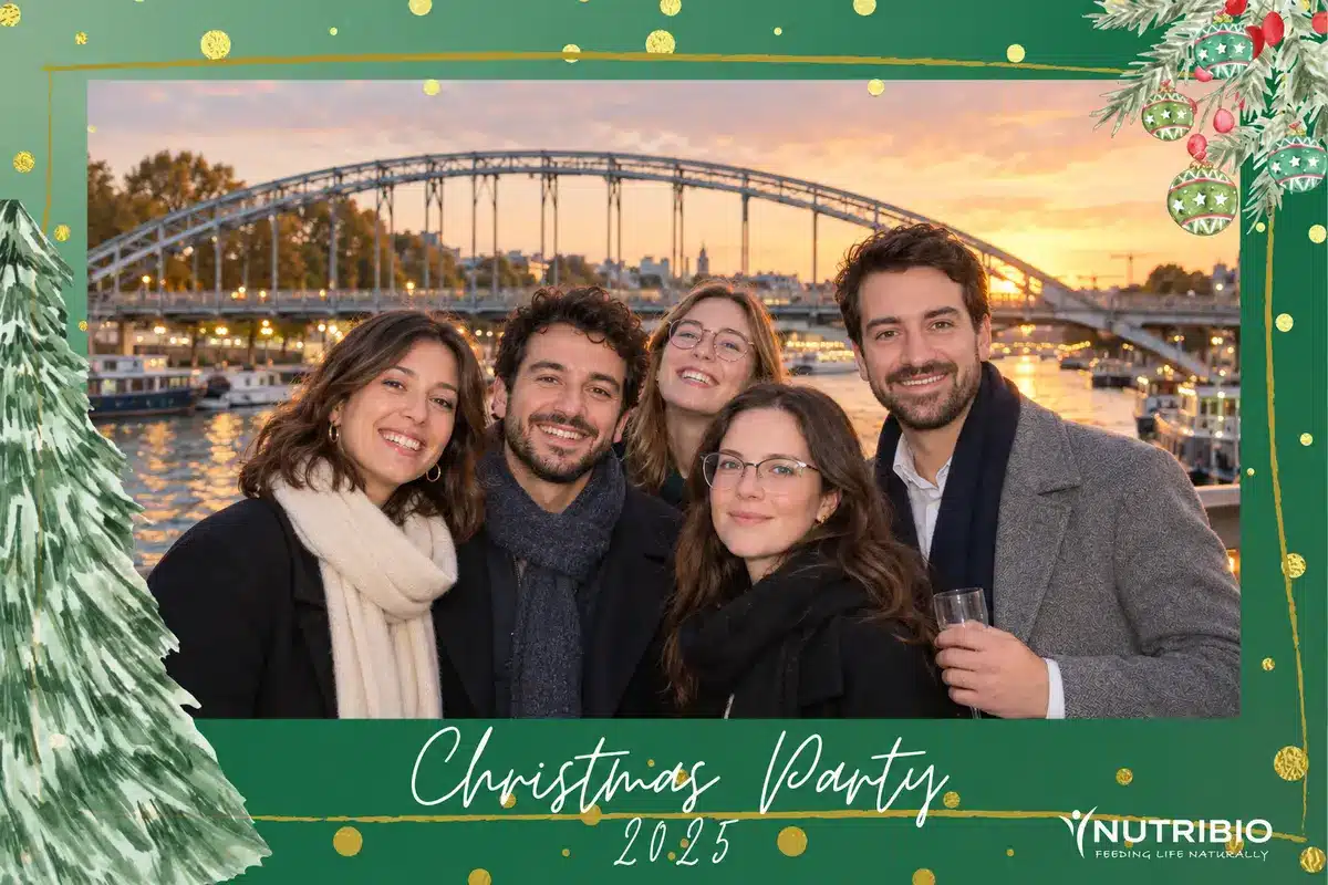 Tirage photobooth de la Christmas Party 2025 de Nutribio : six collègues posent avec la Seine et un pont métallique au coucher du soleil en arrière-plan, cadre vert de Noël