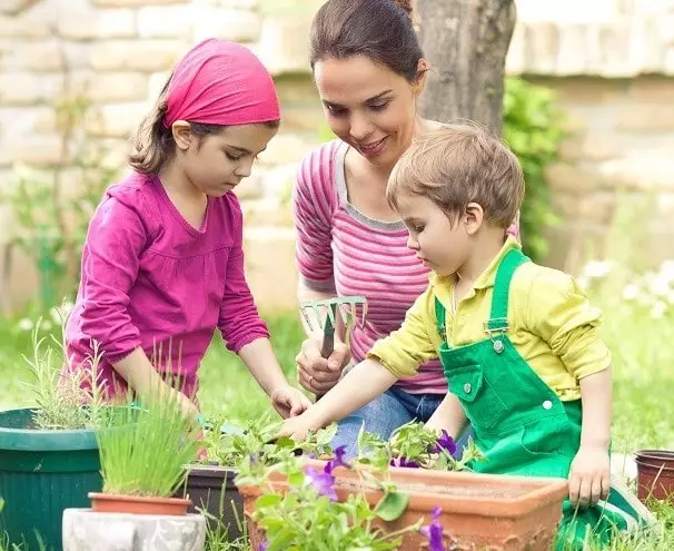 deux enfants et leur maman sont dans le jardin en train de planter fleur et legume dans bac à fleurs