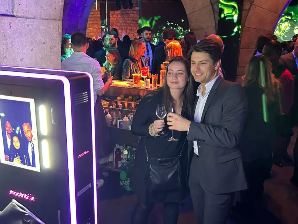 une femme et un homme se prennent en photo un verre à la main devant une borne photo selfie lors d'une soirée dans un bar avec barman faisant des cocktail en fond